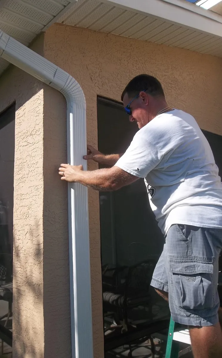 Installer fastening a white downspout to a stucco wall beside a screened lanai