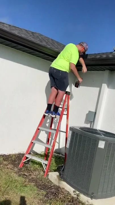 Installer on a ladder fitting a water diverter at the roof edge above an AC unit